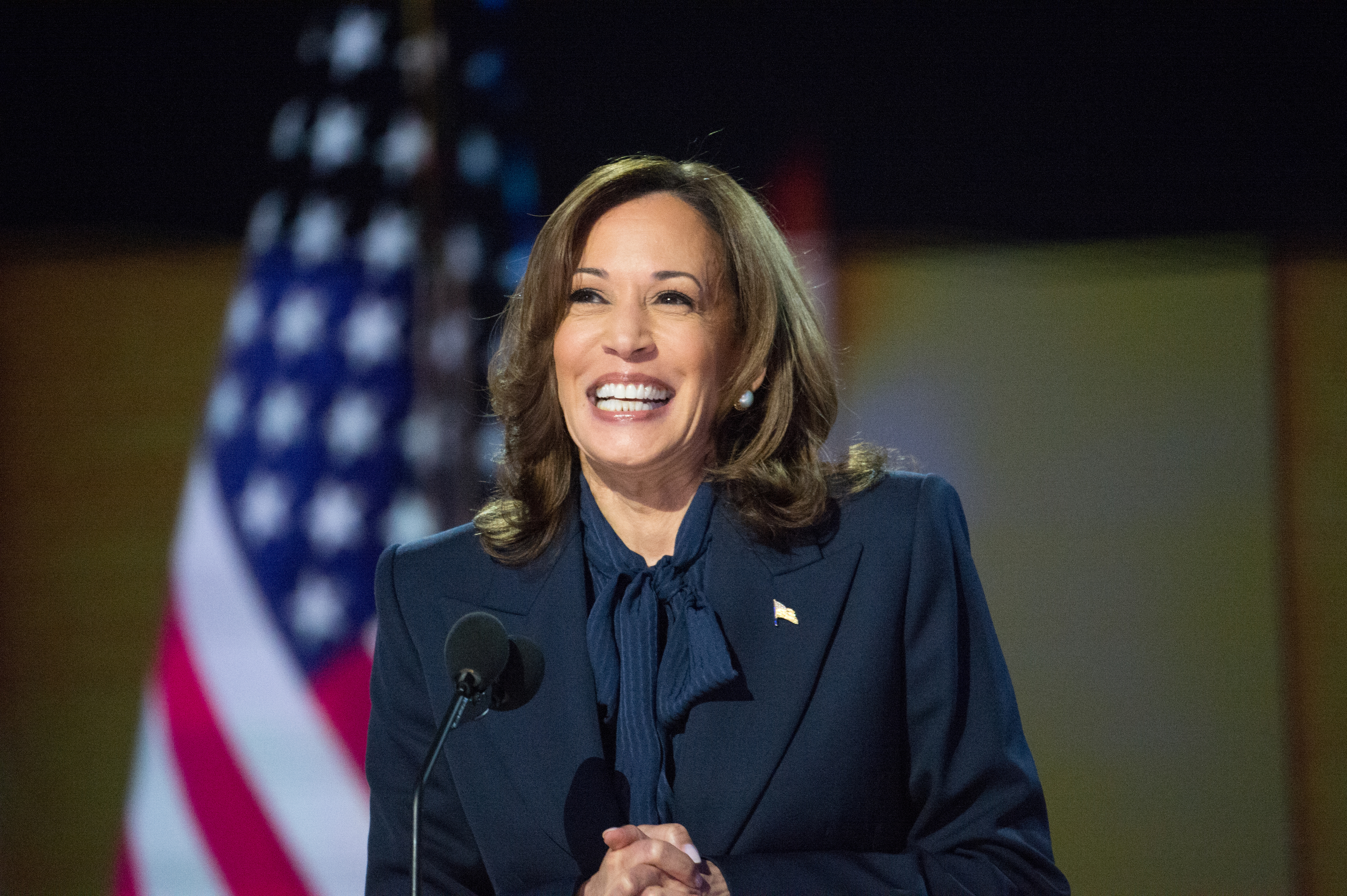 Kamala Harris smiles while speaking at a podium with an American flag in the background. She is wearing a navy blazer and a matching blouse with a bow