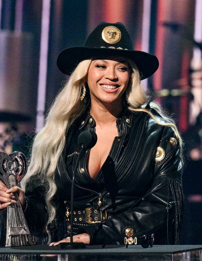 Beyoncé wears a black, Western-inspired outfit with a wide-brimmed hat, smiling while holding an award and standing at a microphone onstage