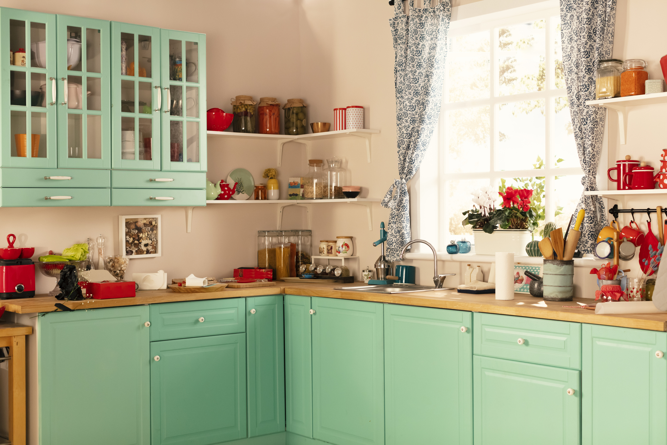 A cozy, well-lit kitchen with mint-green cabinets, wooden countertops, a sink under a window with floral curtains, and shelves holding various jars and kitchen items