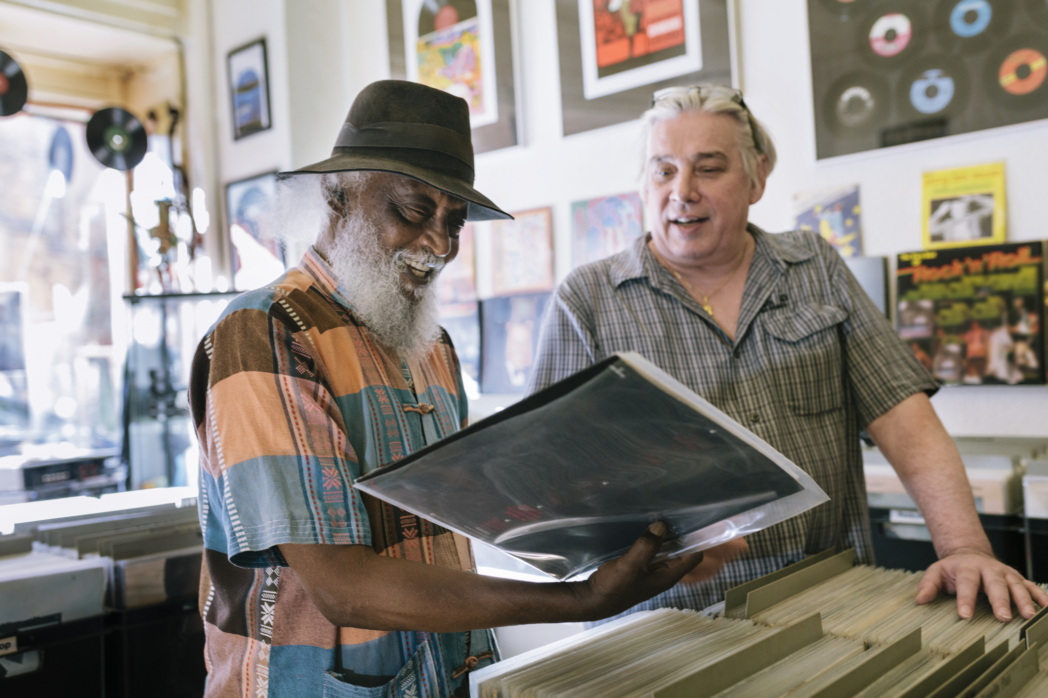 Two men in a record shop looking at vinyl records together; one wears a colorful shirt and hat, the other a plaid shirt. Various records and posters are visible