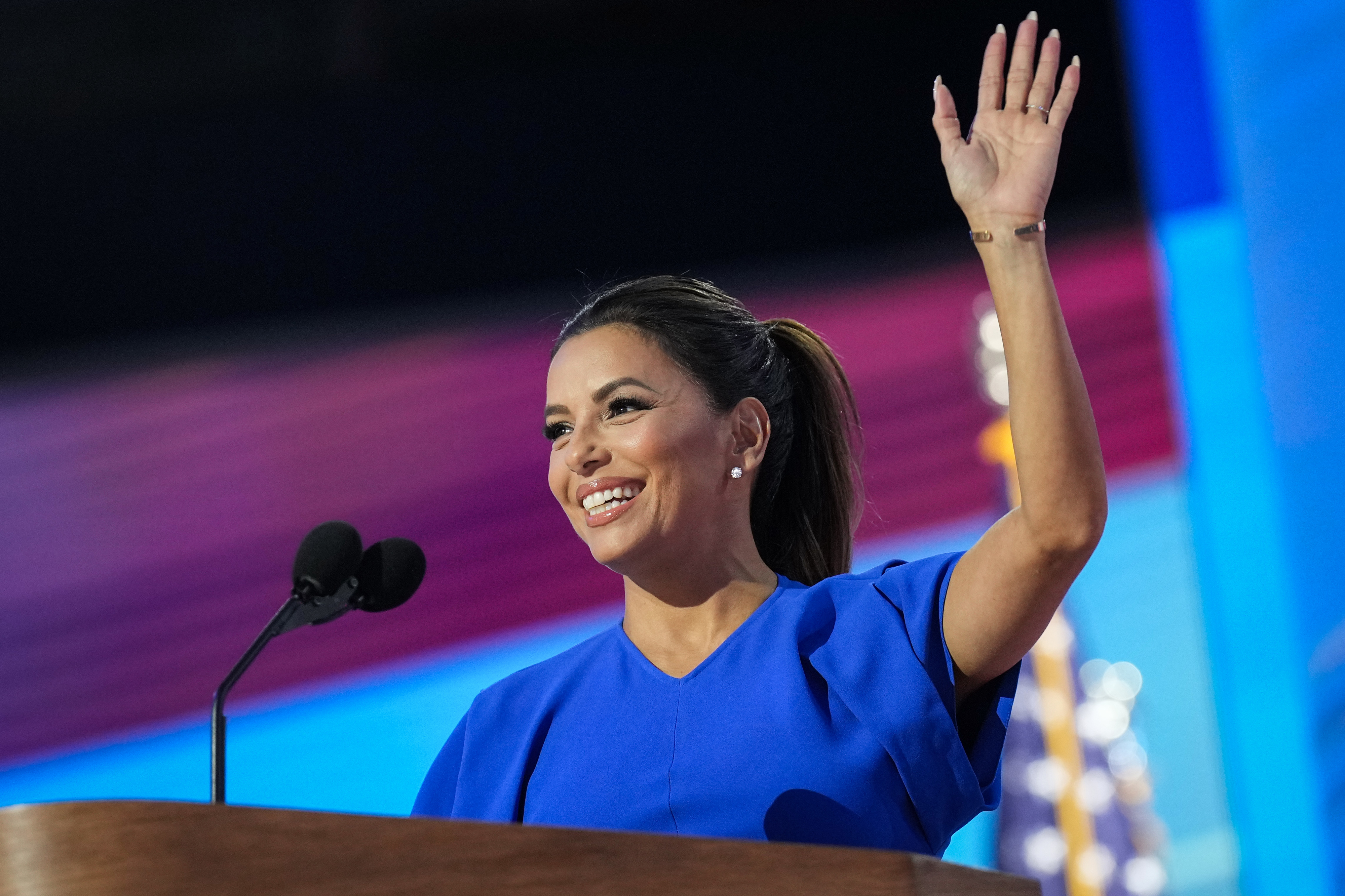 Eva Longoria smiles and waves while speaking at a podium. She is wearing a simple, elegant dress
