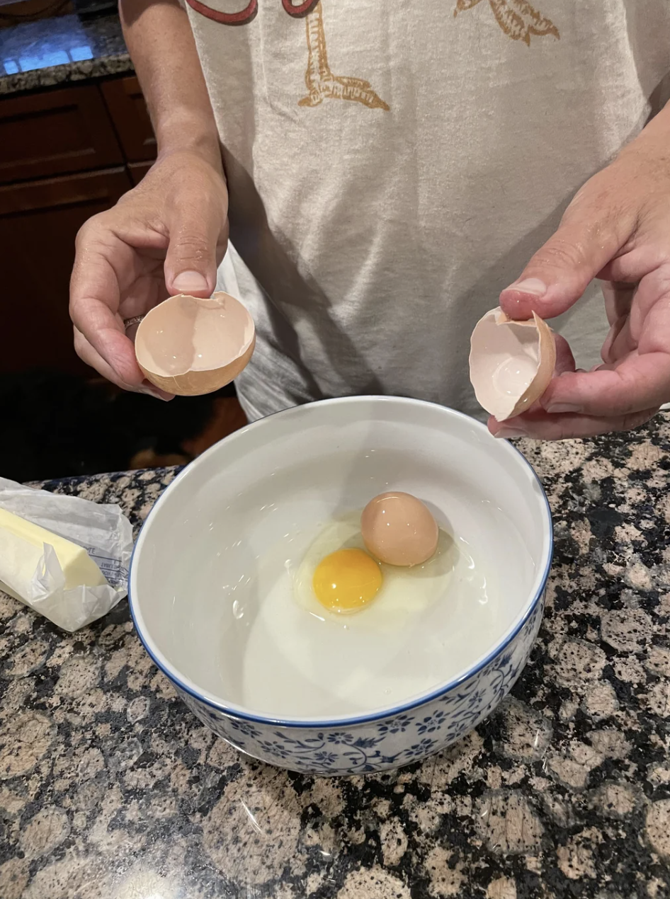 A person is cracking an egg into a bowl on a granite countertop. One egg is already in the bowl, and they hold an empty eggshell in each hand