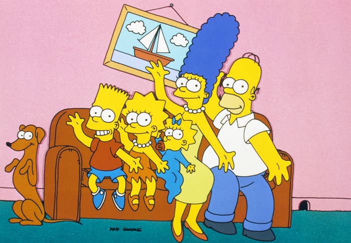The Simpsons family, including Bart, Lisa, Maggie, Marge, and Homer, sit on a couch with their dog, Santa's Little Helper. They pose with a picture of a sailboat