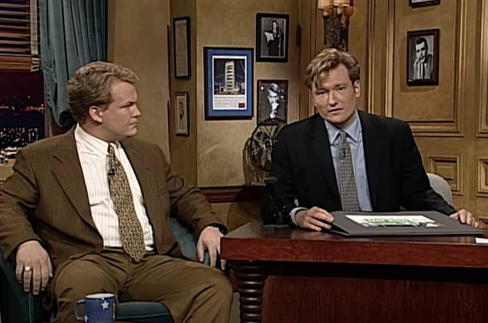Conan O'Brien and Andy Richter sitting at a desk on a late-night talk show set, engaged in conversation