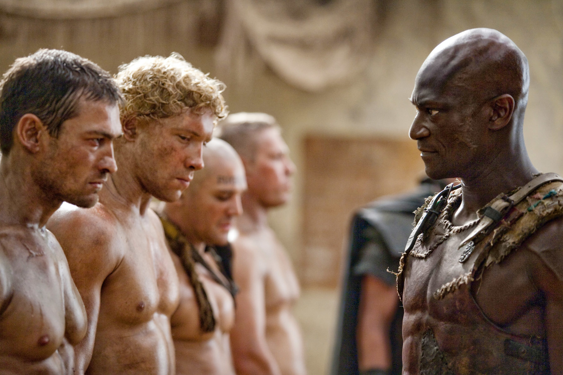 Four shirtless men with serious expressions stand in a line facing a fifth shirtless man who is also serious. Scene suggests a confrontation