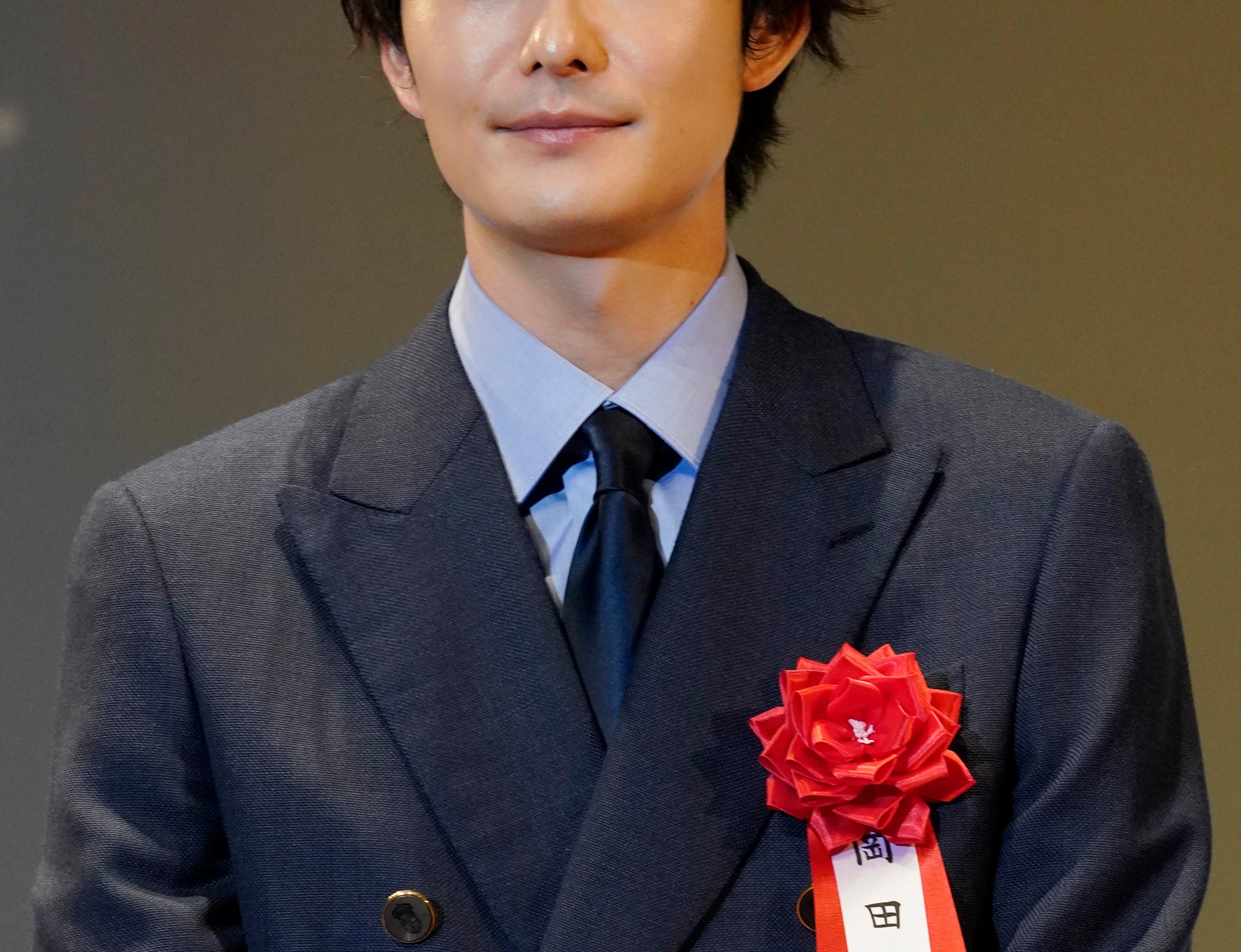 男性がダブルのスーツを着ており、左胸には赤いリボンのついた名札がついている。