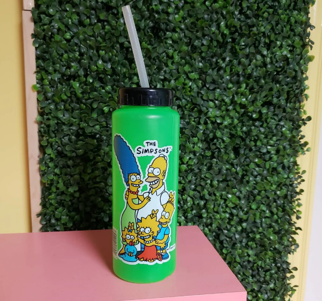 A green water bottle featuring characters from The Simpsons: Homer, Marge, Bart, Lisa, and Maggie. It stands on a pink surface against a green leafy background