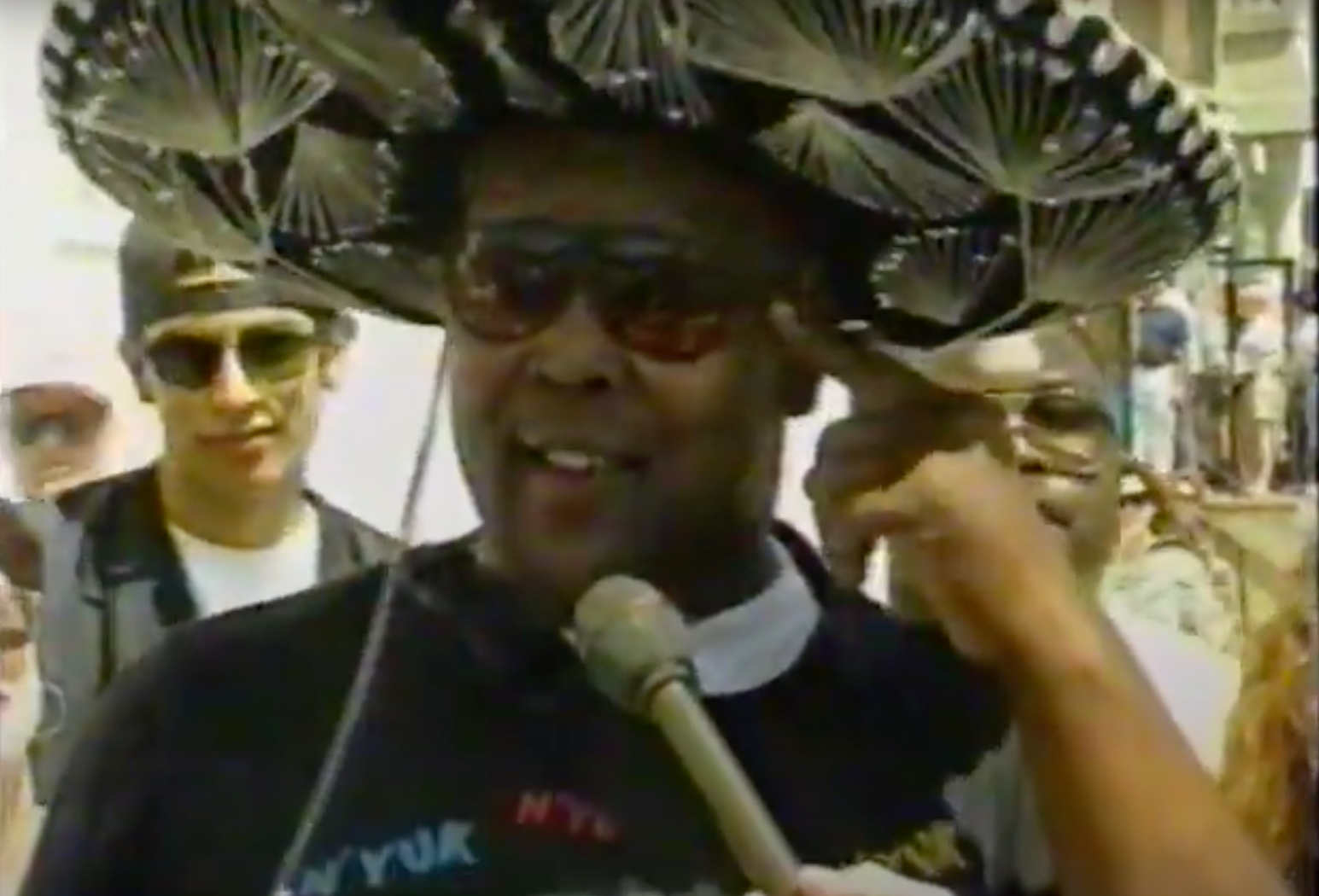Man wearing a large, decorative hat and sunglasses speaks into a microphone while others stand behind him, some also wearing sunglasses