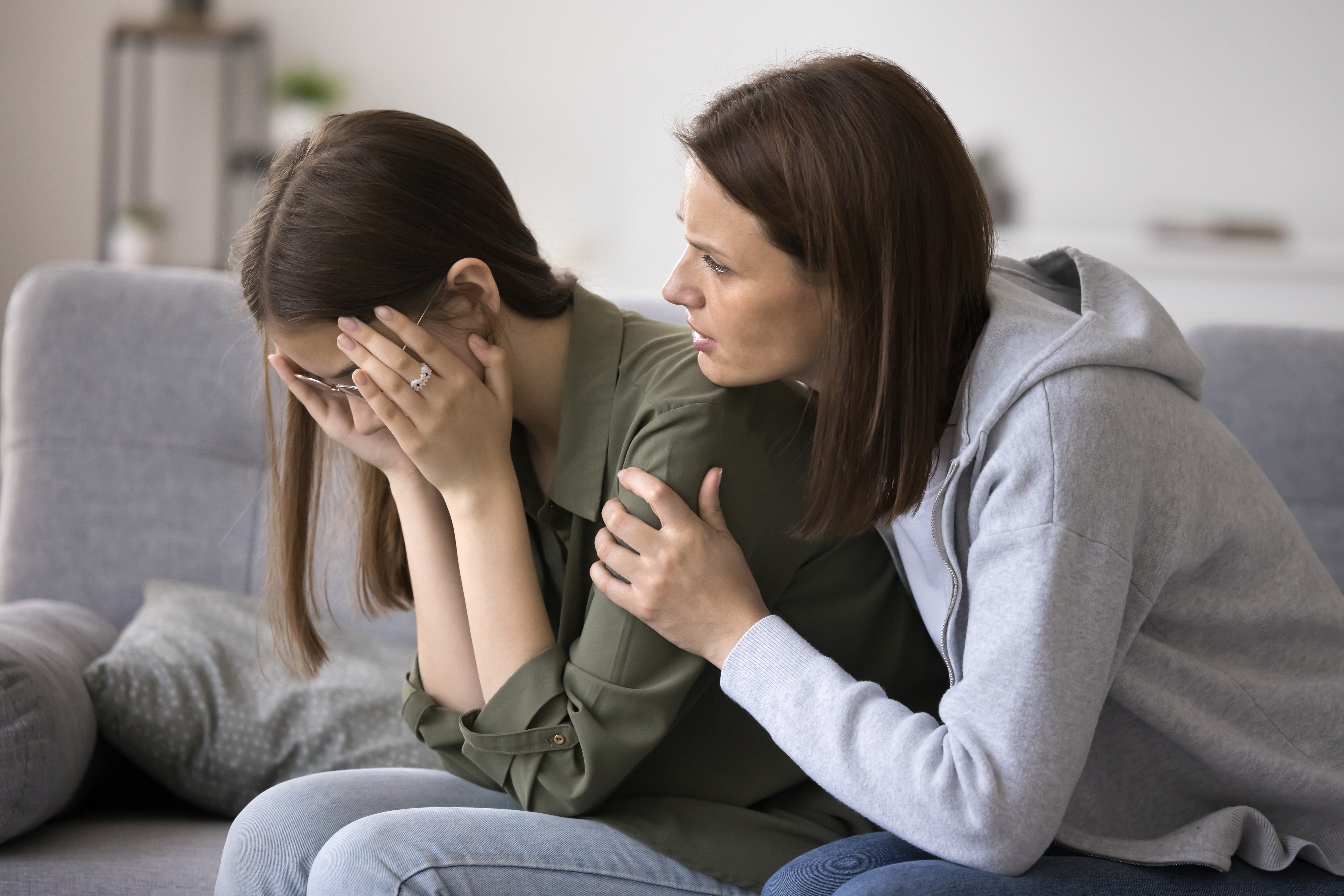 A woman wearing a gray sweater sits on a couch comforting a younger girl in a green shirt who is covering her face with her hands