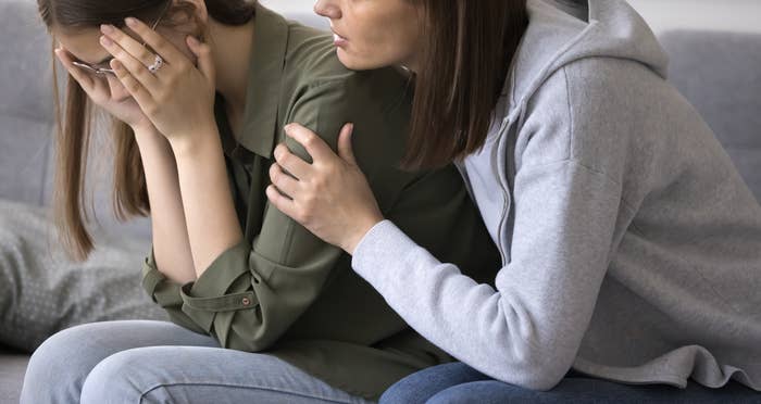 A woman wearing a gray sweater sits on a couch comforting a younger girl in a green shirt who is covering her face with her hands