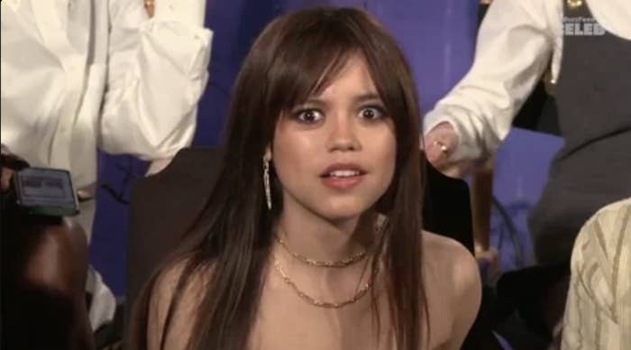 Jenna Ortega looks surprised at an event. She is wearing a strapless top with a gold necklace and earrings. Other people are visible in the background