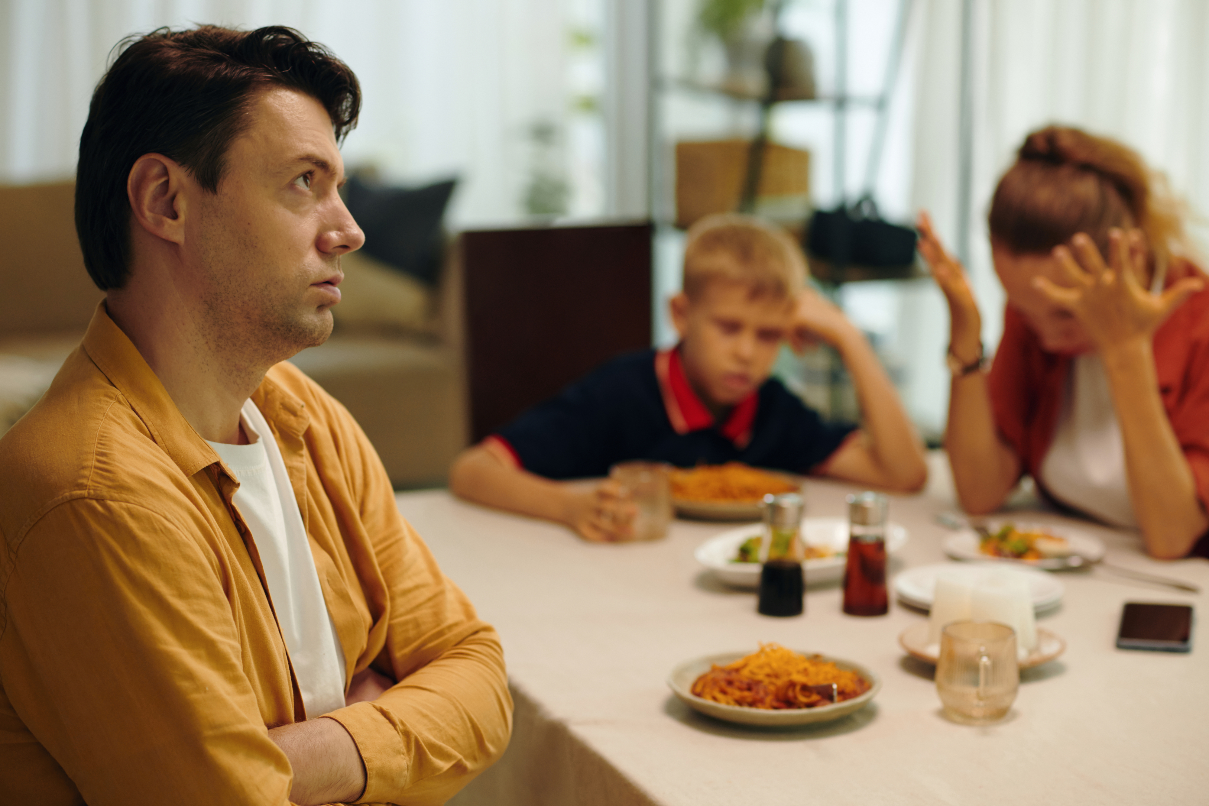 An upset man sits with crossed arms at a dining table, while a woman, holding her head in frustration, and a sad boy sit nearby with food in front of them