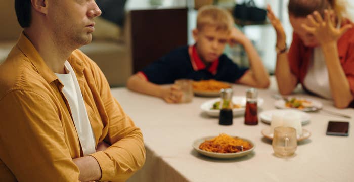 An upset man sits with crossed arms at a dining table, while a woman, holding her head in frustration, and a sad boy sit nearby with food in front of them