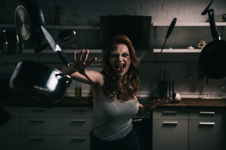 A woman in a tank top and jeans, appearing to use telekinesis to levitate kitchen utensils in a dimly lit kitchen