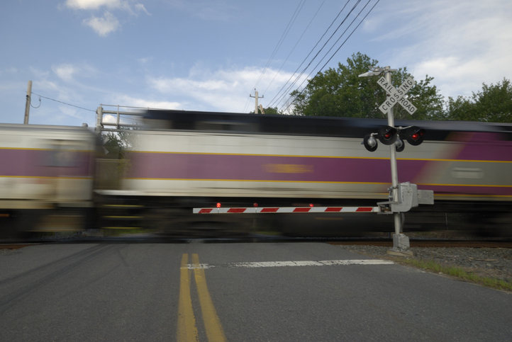 Two trains passing through a railroad crossing, one stationary and one in motion, with a visible crossing sign and barrier. No identifiable people