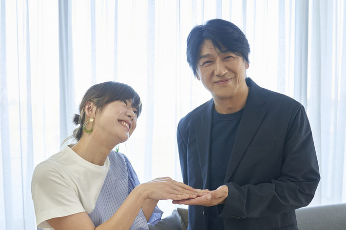 女性と男性が室内で微笑んでいる。女性は白いTシャツとストライプのワンピースを着用し、男性は黒いジャケットを着ている。