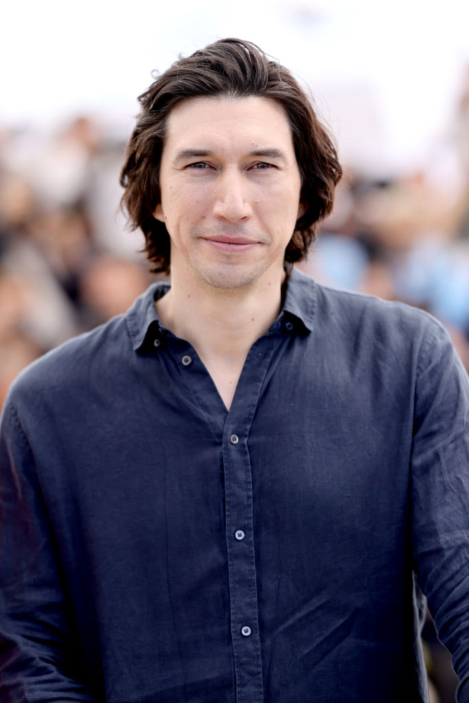 Adam Driver stands facing the camera, wearing a casual button-up shirt