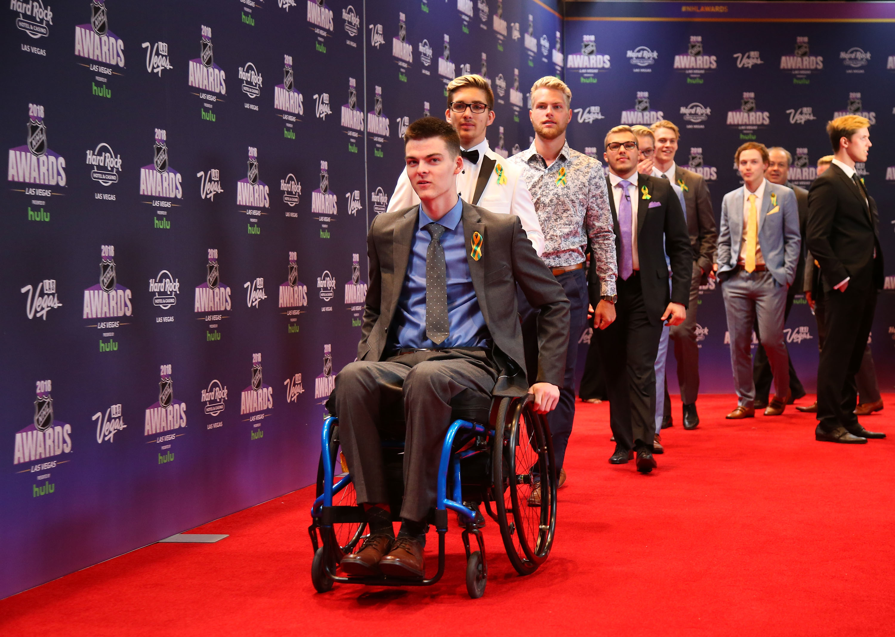 Line of men on a red carpet, led by a man in a blue wheelchair. They are dressed in suits, some with patterned shirts