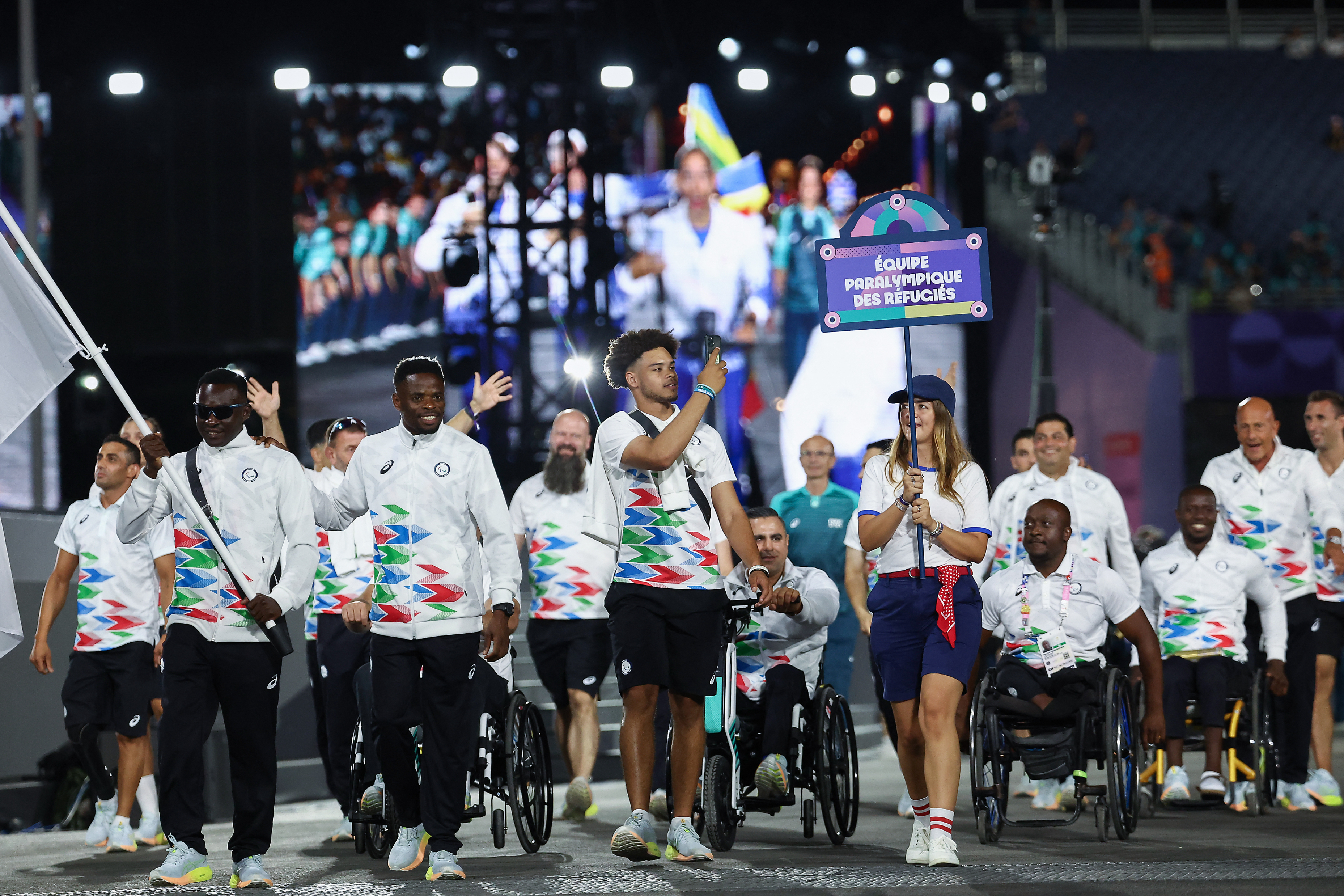 Athletes from the Refugee Paralympic Team march in the opening ceremony, wearing white jackets with colorful patterns. One athlete carries the team's flag