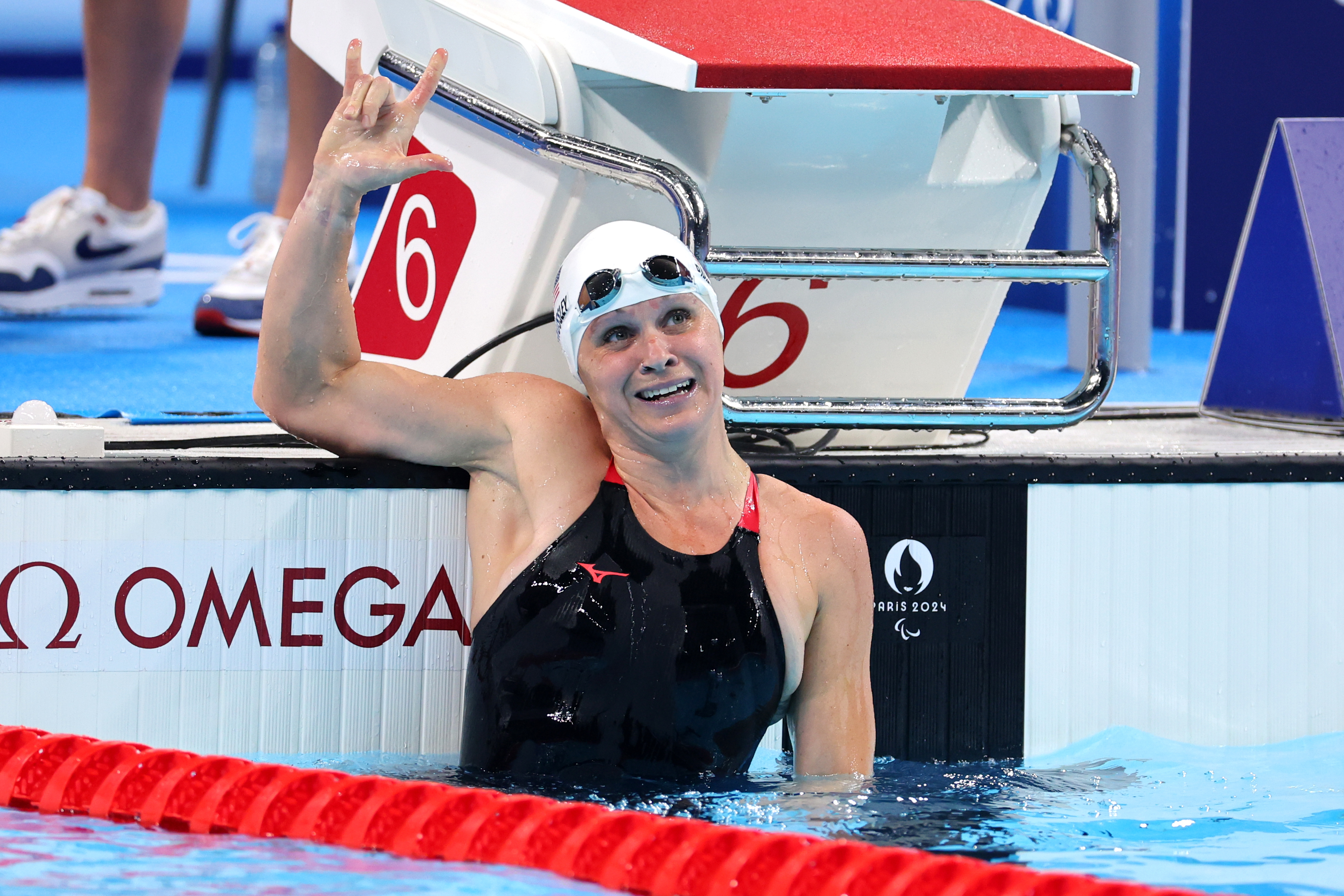 Swimmer with swim cap and goggles, wearing a black swimsuit, celebrates by holding up three fingers at a pool's edge with lane number 6 in the background