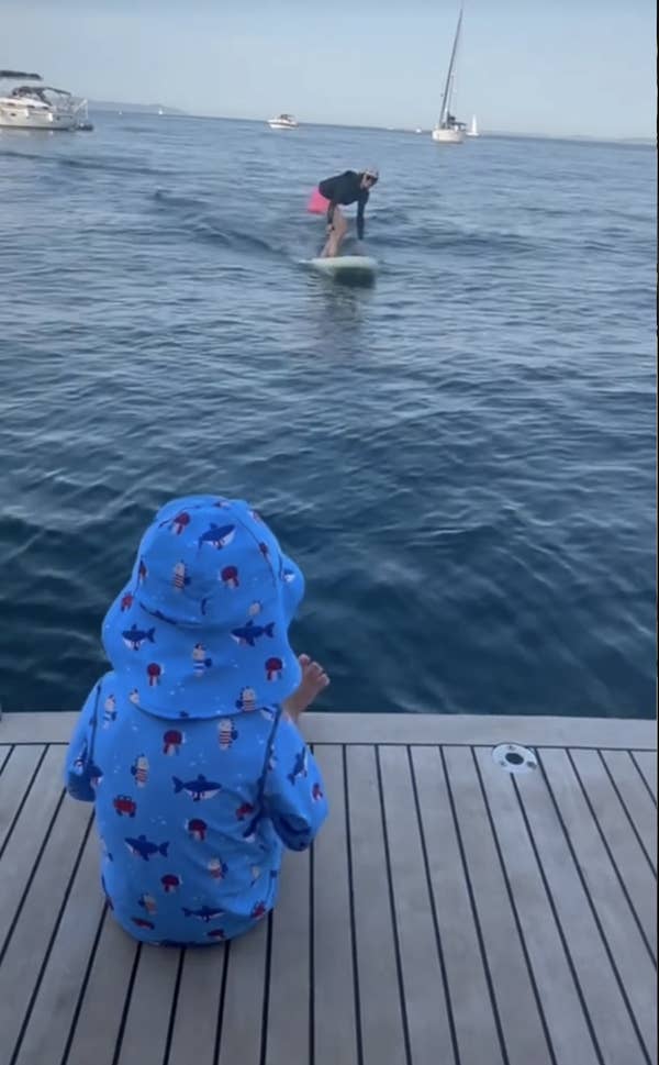 A child in a blue hoodie with a hood stands on a wooden dock, watching an adult paddleboarding on a calm sea. Several boats are in the background