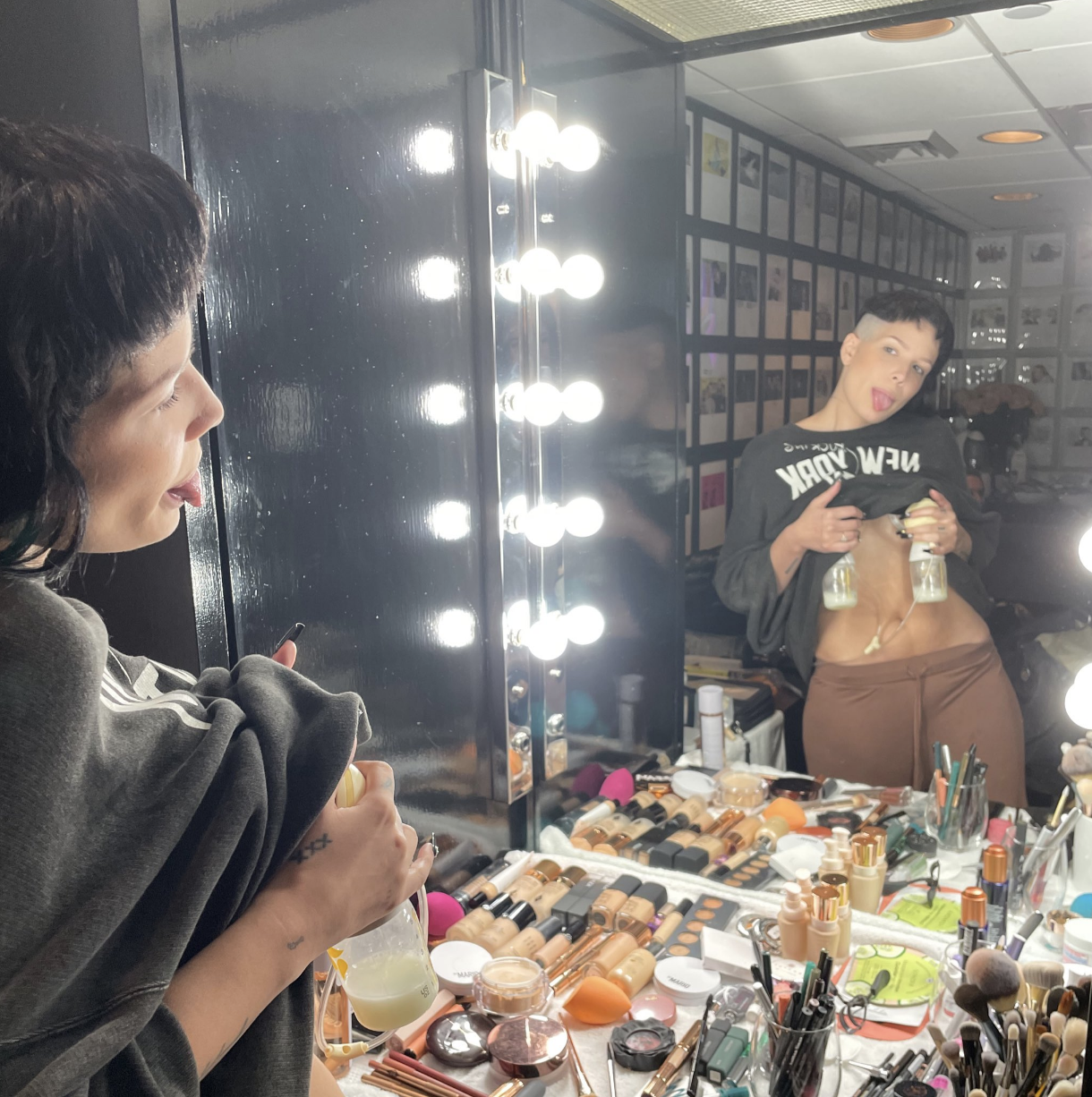 Halsey backstage in a dressing room, wearing a casual outfit and playfully sticking out their tongue in the mirror. The vanity is filled with makeup products