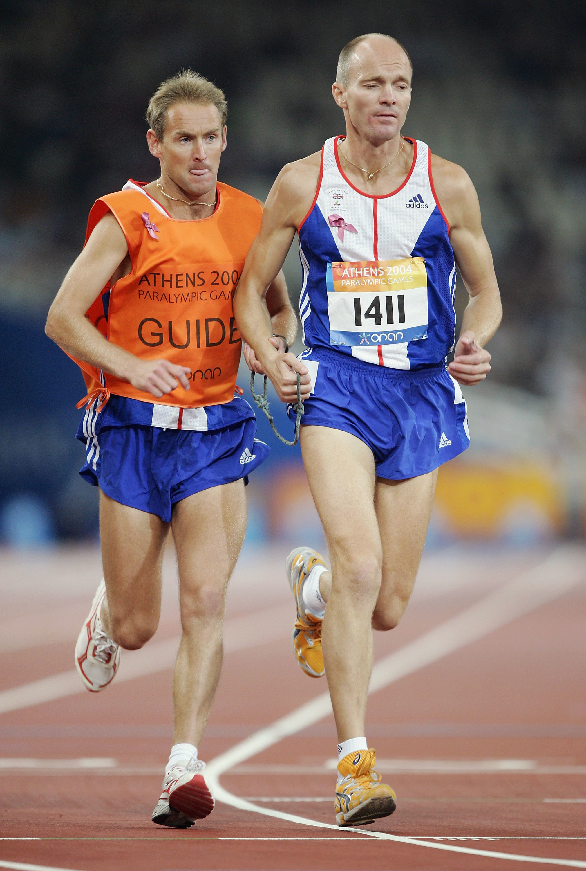 Two athletes, one with a &quot;Guide&quot; bib, and the other with bib number 1411, running on a track at the Athens 2004 Paralympic Games