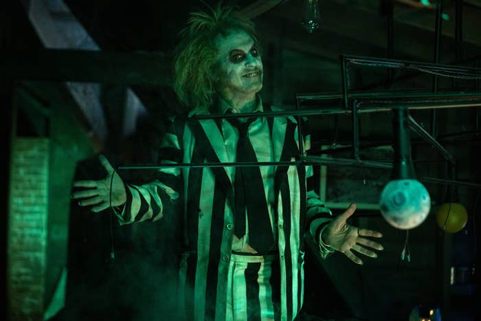 Michael Keaton as Beetlejuice is standing with arms open, wearing a black-and-white striped suit, dark tie, and wild hair, with theatrical makeup on their face