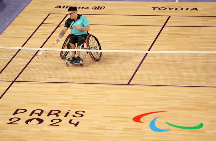 Para-athlete in a wheelchair playing badminton at a Paris 2024 Paralympic event. Logos for Allianz, Toyota, and the Paralympic Games are visible on the court
