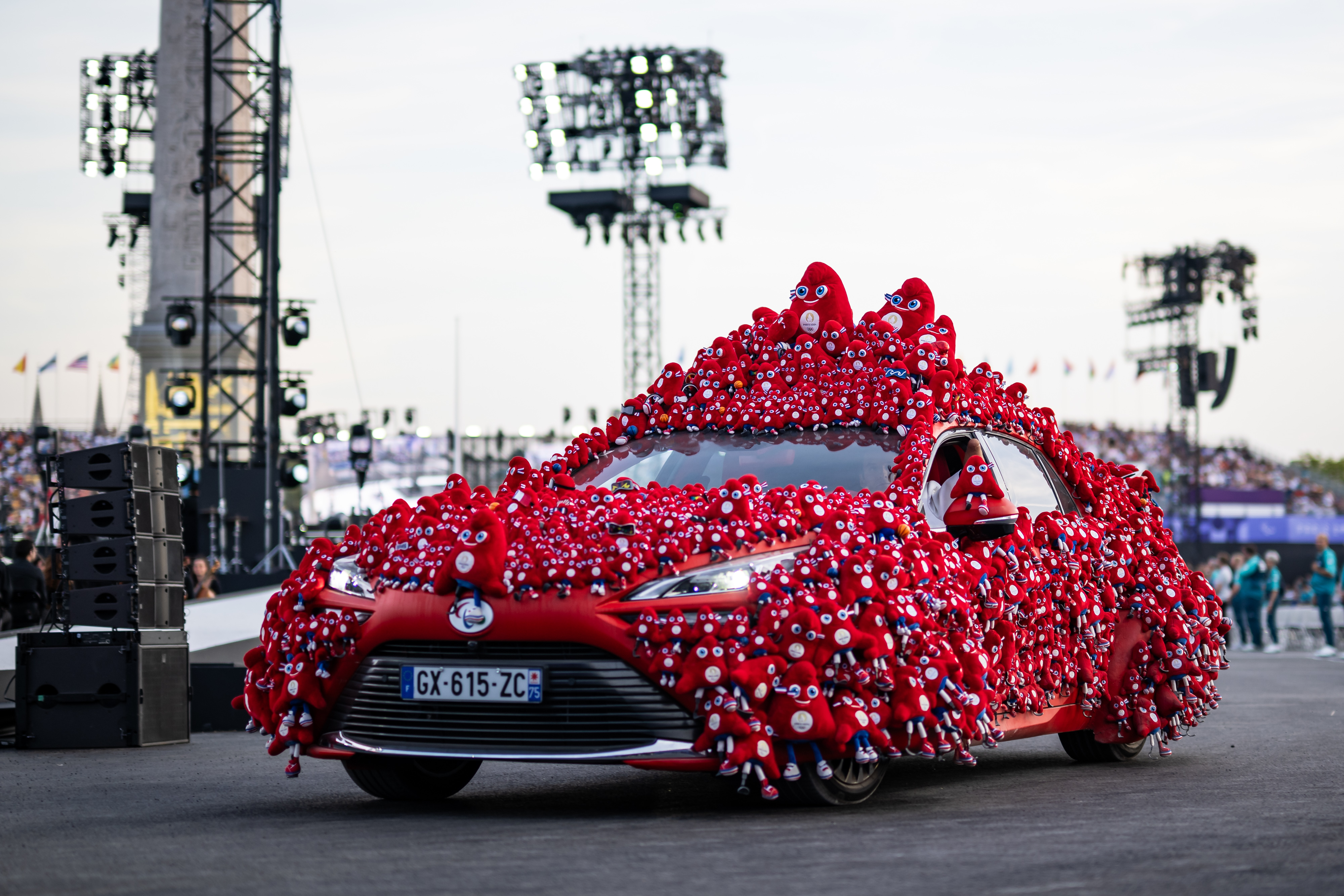 A car covered in numerous plush toy mascots is displayed at an outdoor event with a crowd in the background