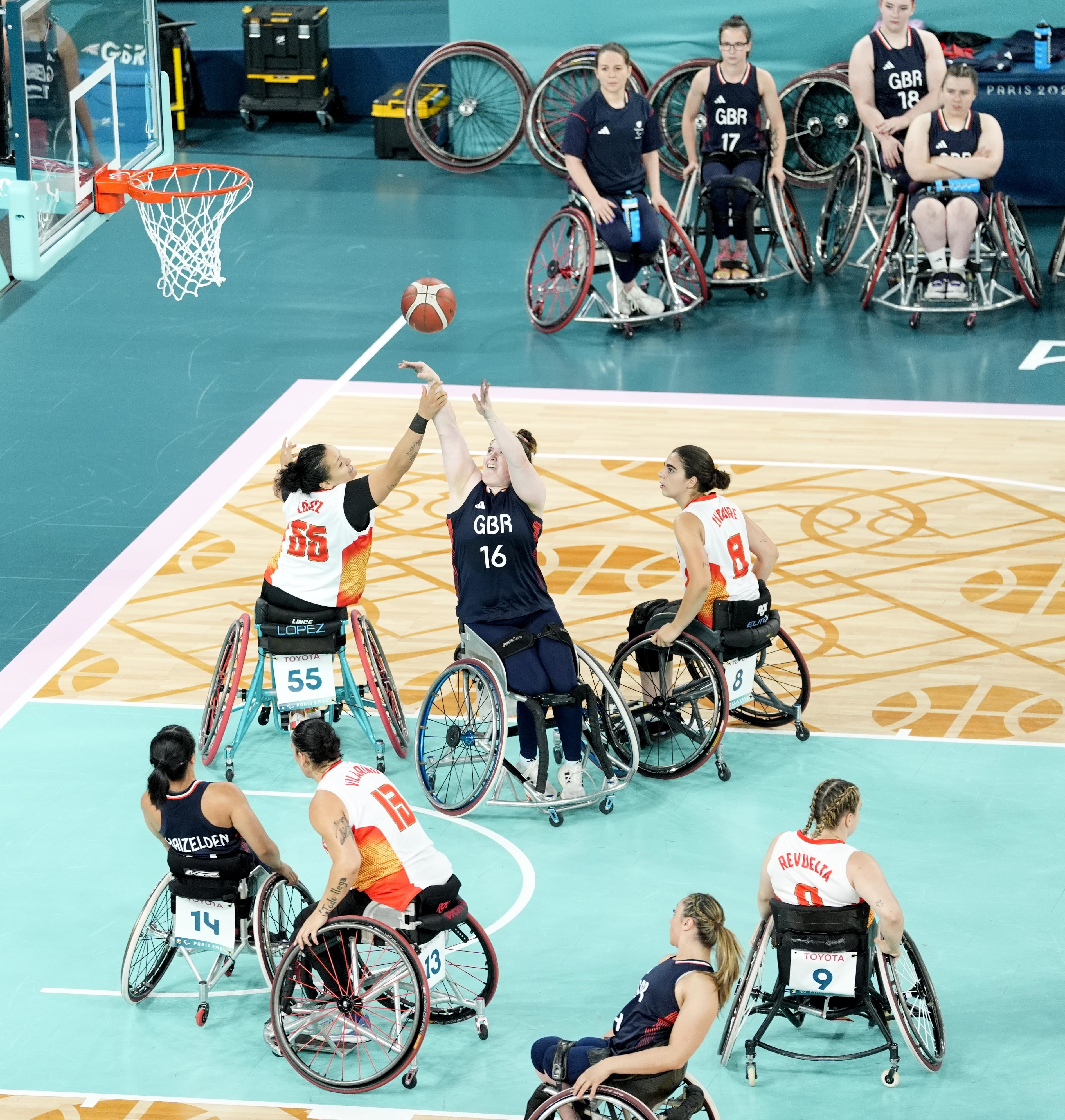 Athletes in wheelchairs compete in a basketball game. Players in blue jerseys from GBR face off against players in white and red jerseys from another team