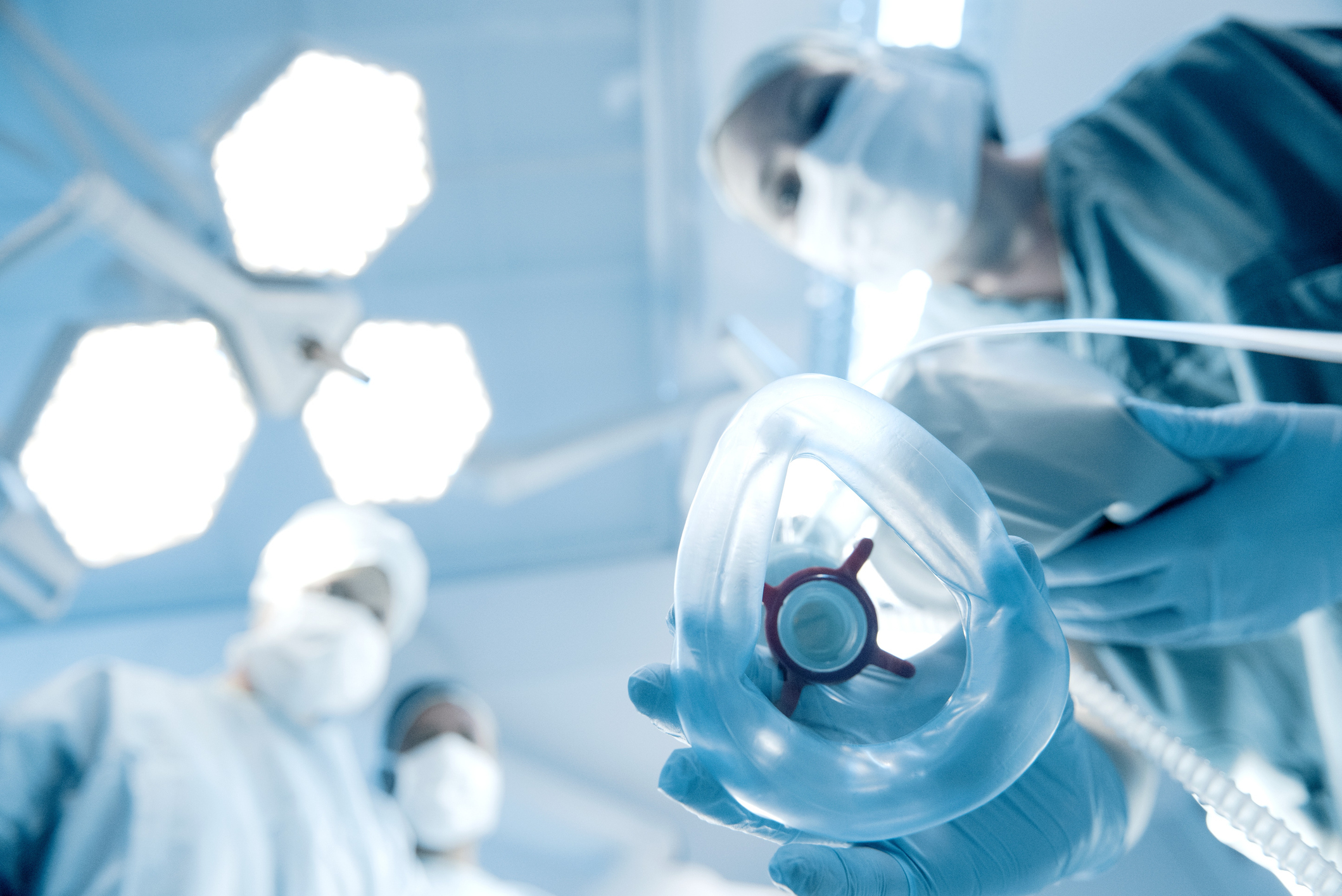 Three medical professionals, wearing surgical masks and scrubs, prepare to administer anesthesia through a mask, viewed from the patient&#x27;s perspective