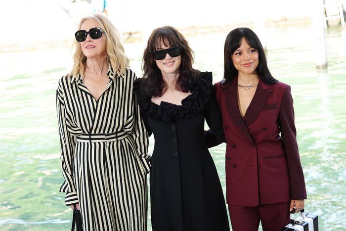 Catharine O'Hara, Winona Ryder, and Jenna Ortega pose together. Catharine wears a striped dress, Winona a black ruffled dress, and Jenna a burgundy suit