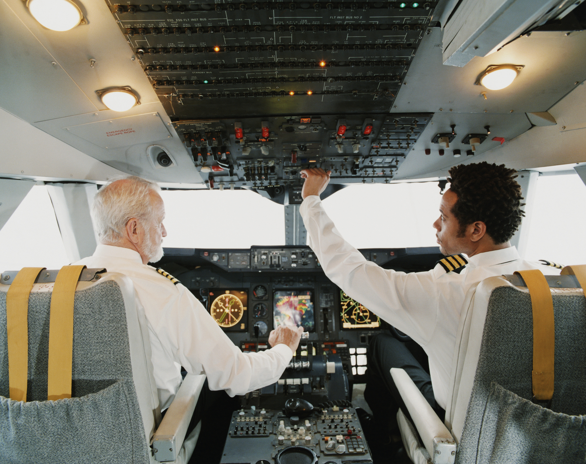 Two pilots in a cockpit are preparing for flight. They are pressing buttons and checking instruments on the control panel