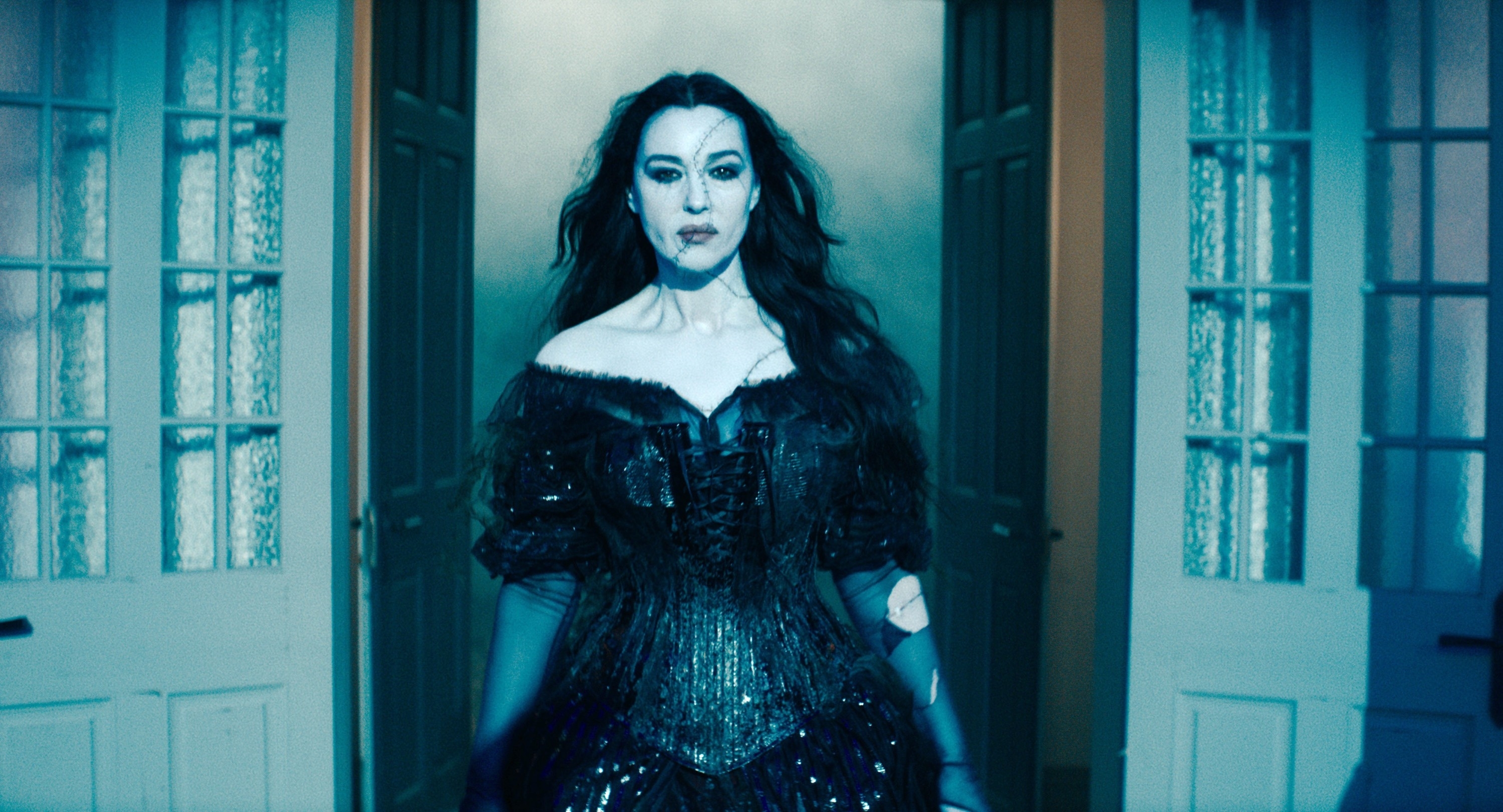 Monica Bellucci stands between two open doors wearing an off-the-shoulder, sequined gown