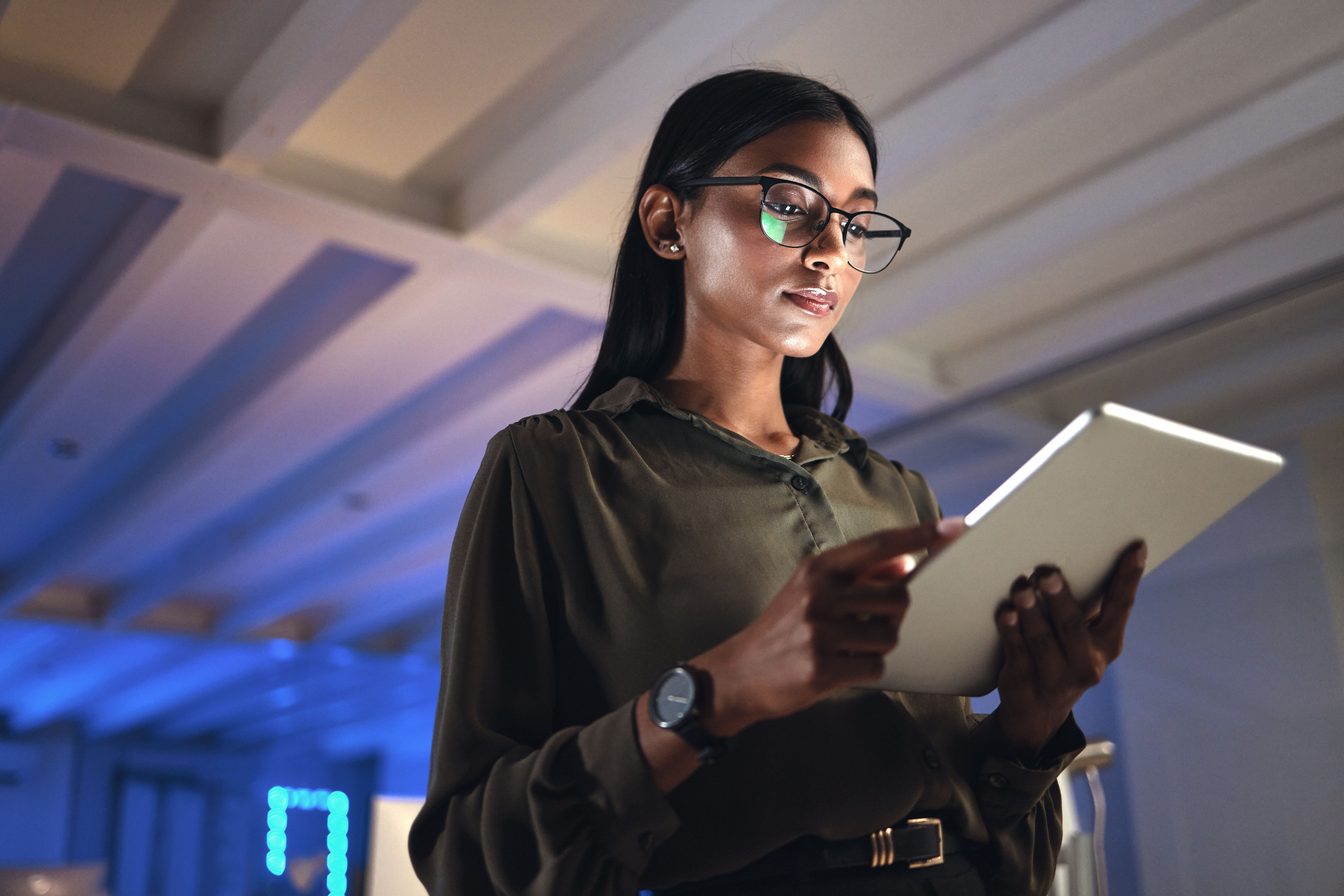A person stands indoors, focused on a tablet. Their attire includes a long-sleeve blouse and glasses. The setting appears to be an office or workspace