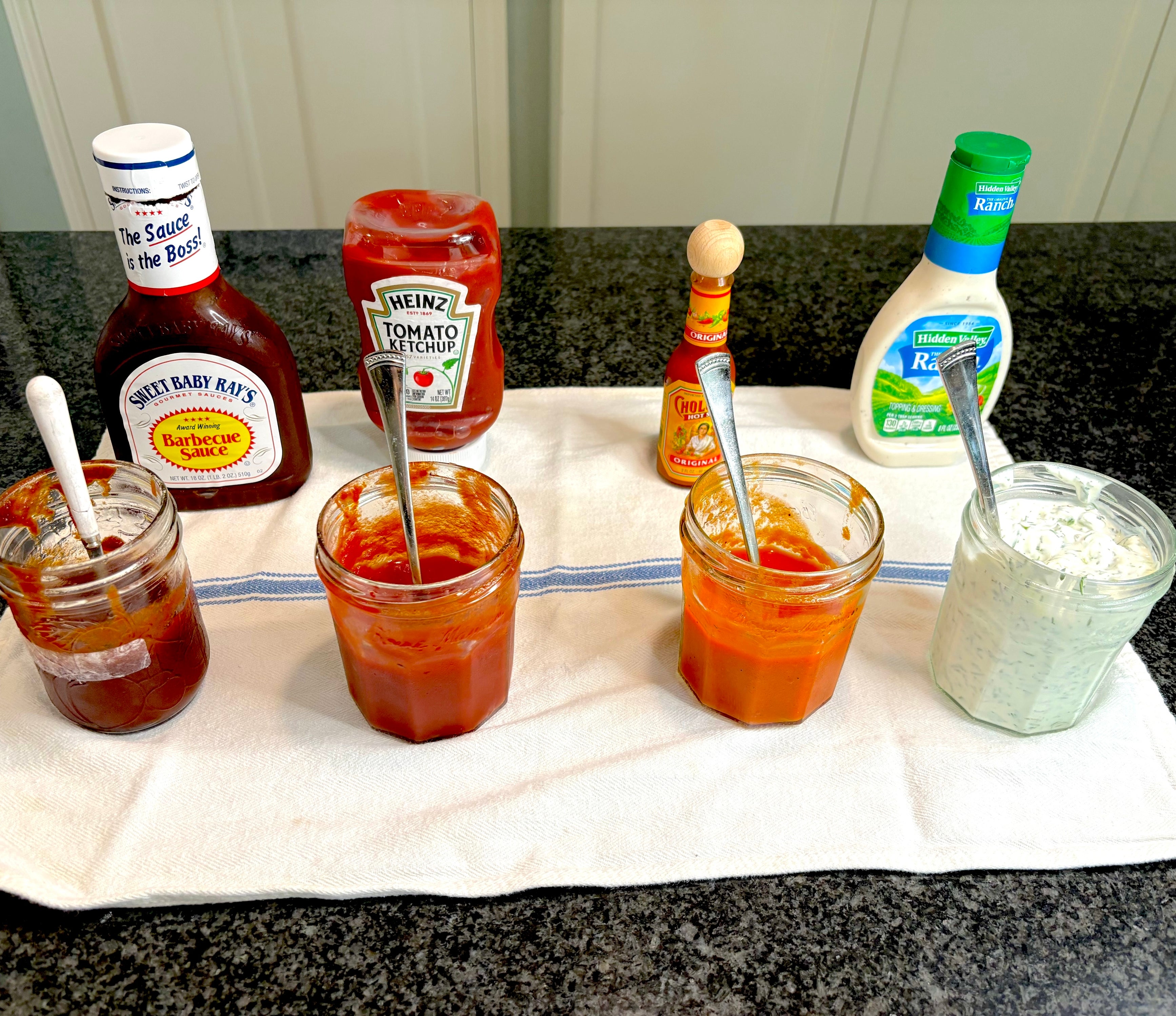 Various condiments, including barbecue sauce, ketchup, hot sauce, and ranch dressing, arranged on a kitchen counter with spoons in small jars