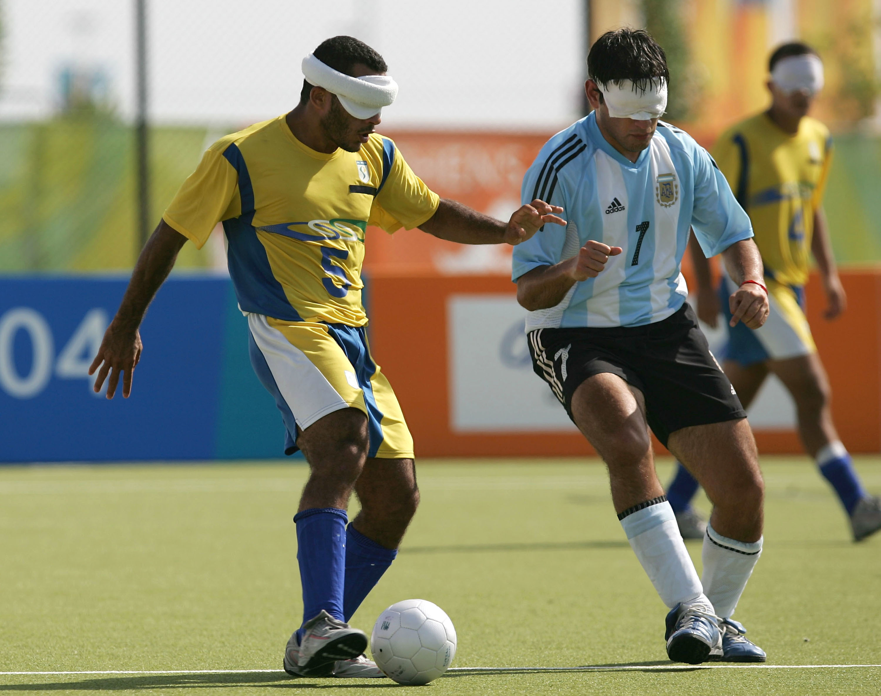 Two blindfolded soccer players, one wearing yellow and blue, the other in a light blue and white jersey, compete for the ball during a match