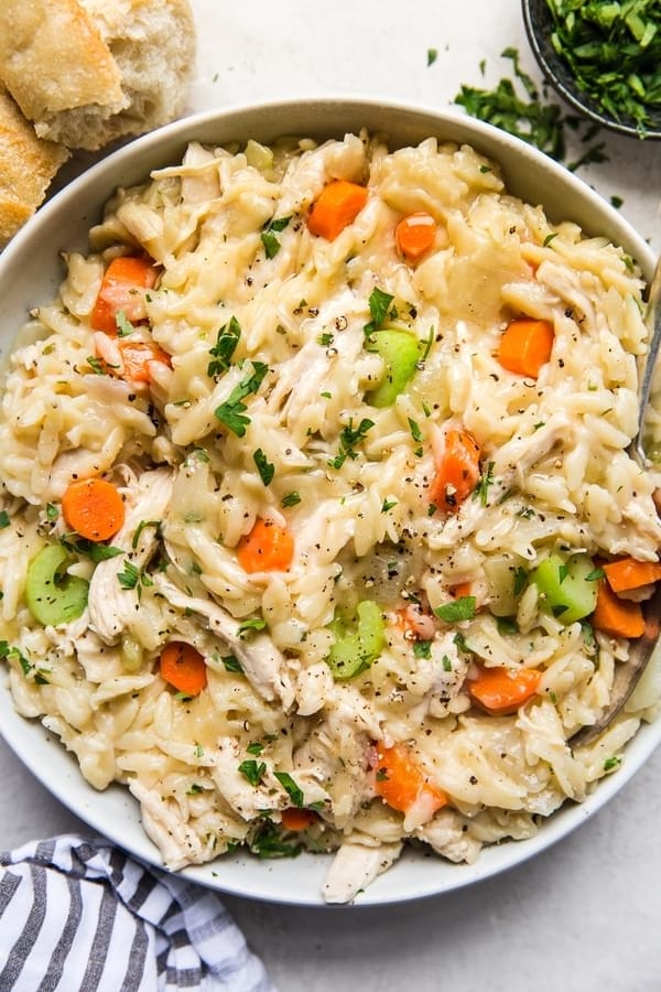 A bowl of creamy chicken and orzo soup garnished with fresh parsley, containing pieces of chicken, carrots, celery, and a creamy broth, with bread on the side