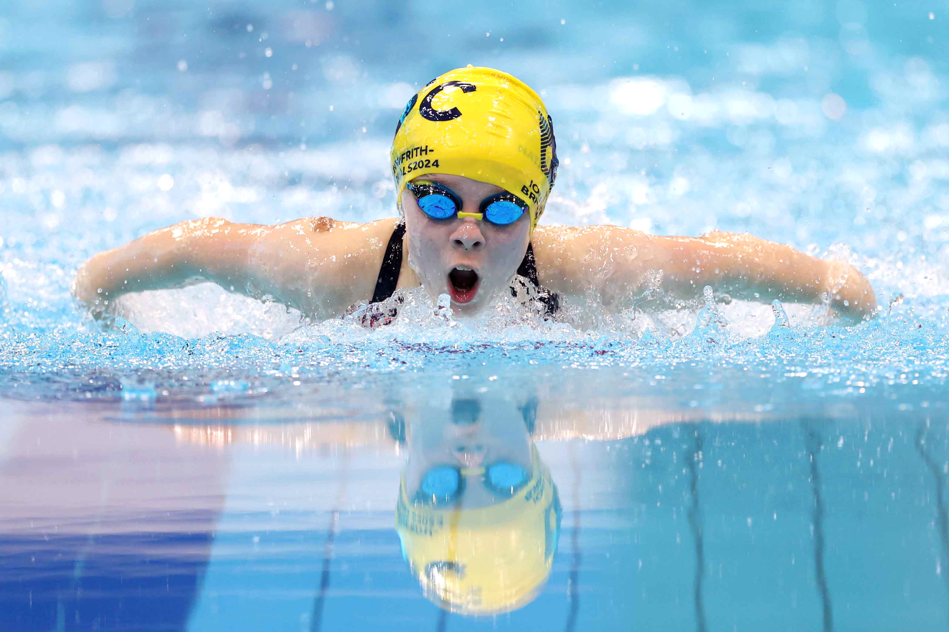 Swimmer during a butterfly stroke in a pool wearing a yellow cap and goggles, with water splashing around