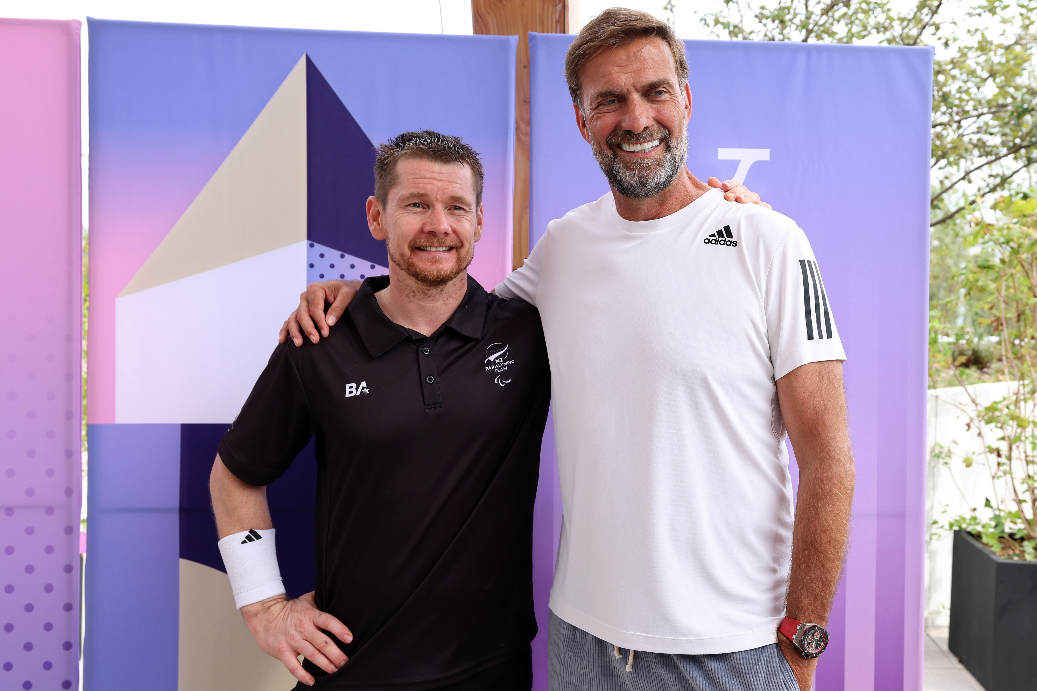 Two men are standing together, smiling. One is wearing a black polo shirt and the other a white athletic shirt