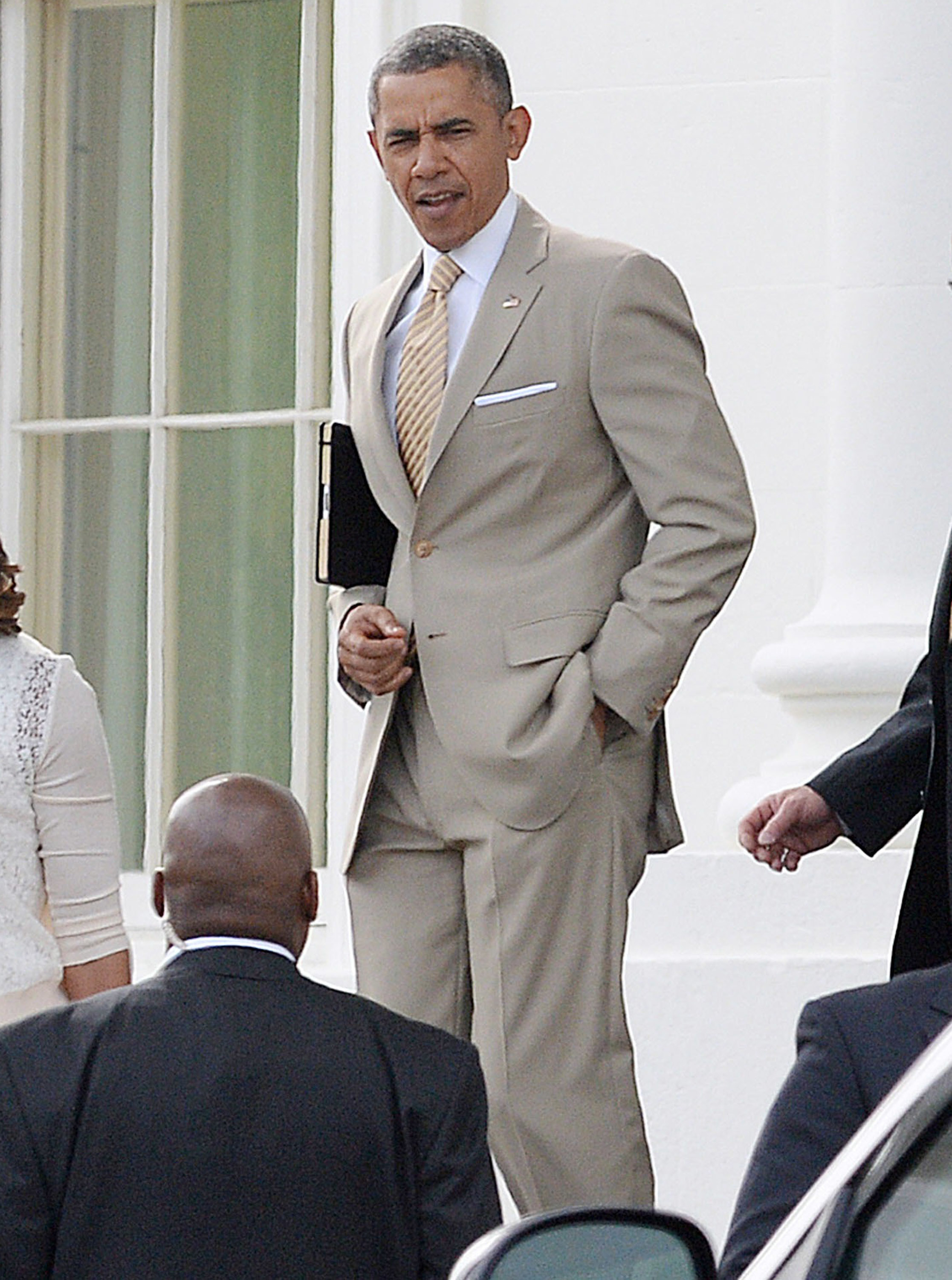Barack Obama is dressed in a beige suit with a striped tie, holding a notebook and talking to people around him