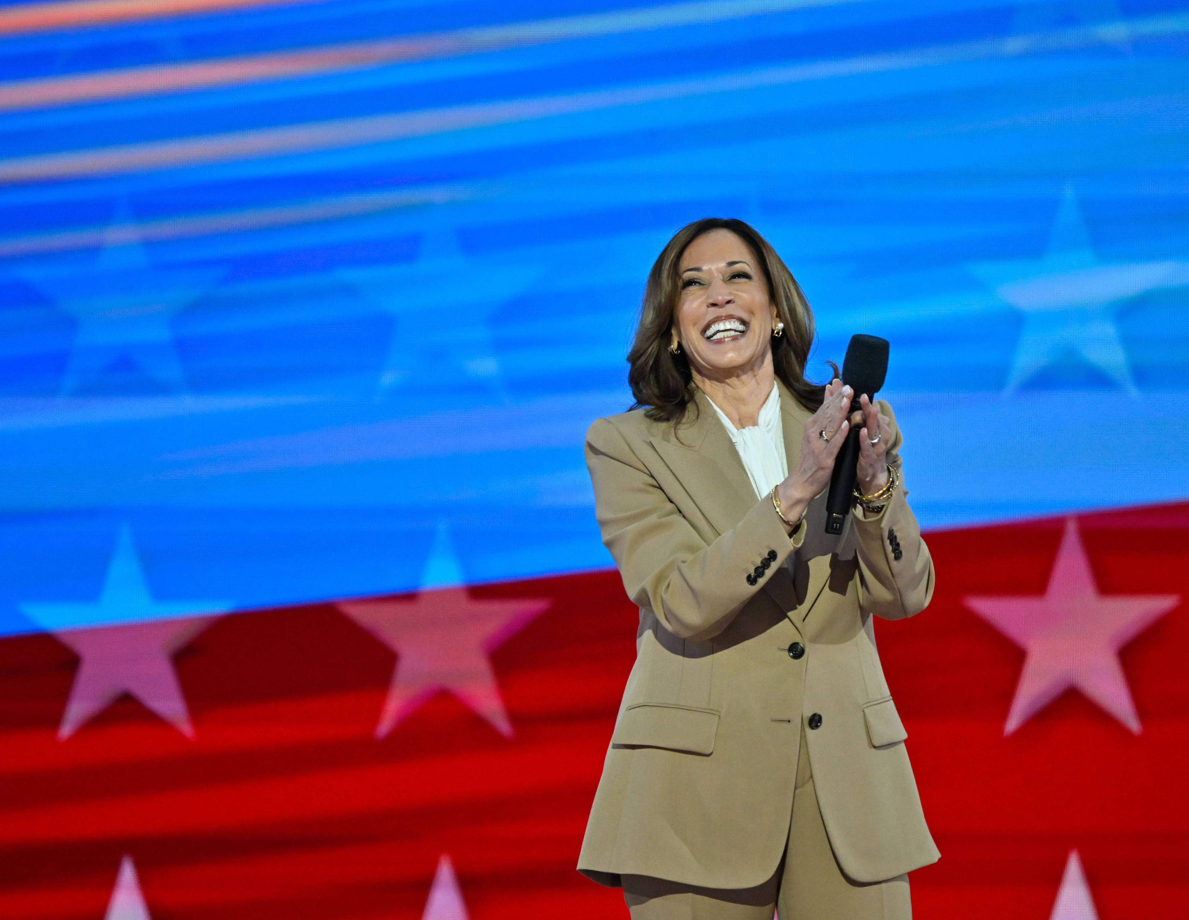 Kamala Harris, smiling and clapping, stands on stage in a tan suit against a backdrop with an American flag design
