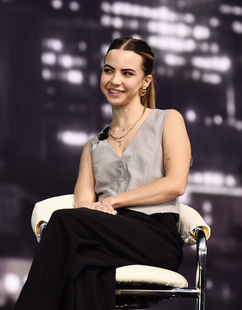 Bobbi Althoff sits in a modern chair, smiling, wearing a sleeveless top and high-waisted pants during a talk show appearance