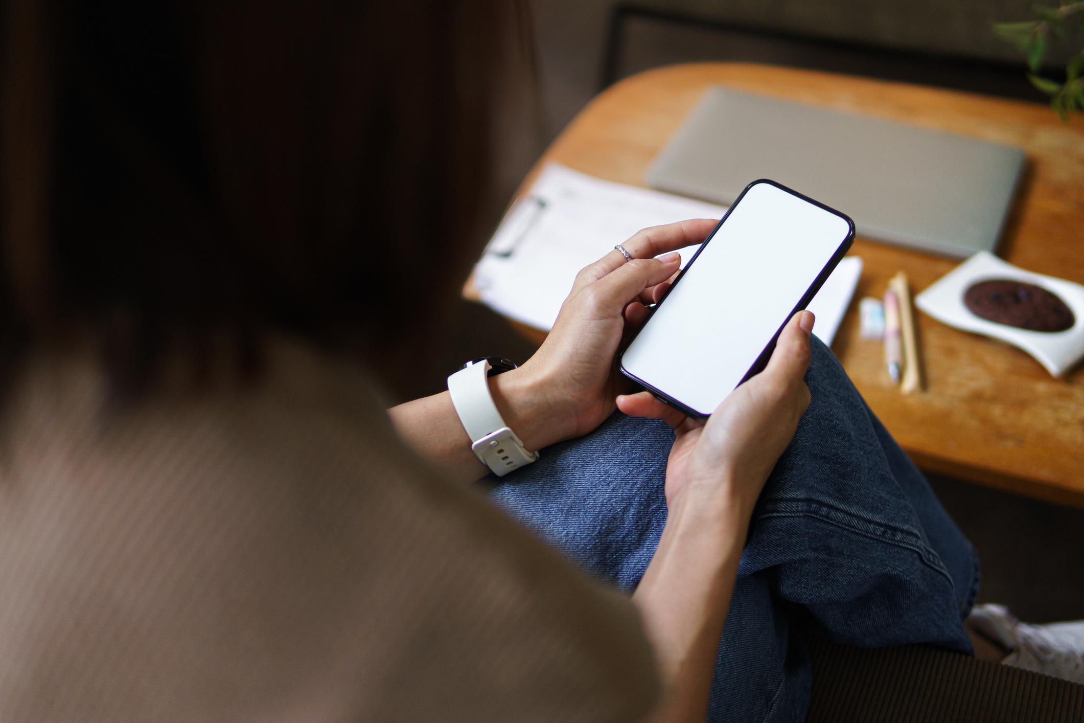 Person wearing casual clothing sits on a couch and looks at a smartphone. A laptop, a notebook, and some pens are on the wooden table in front of them