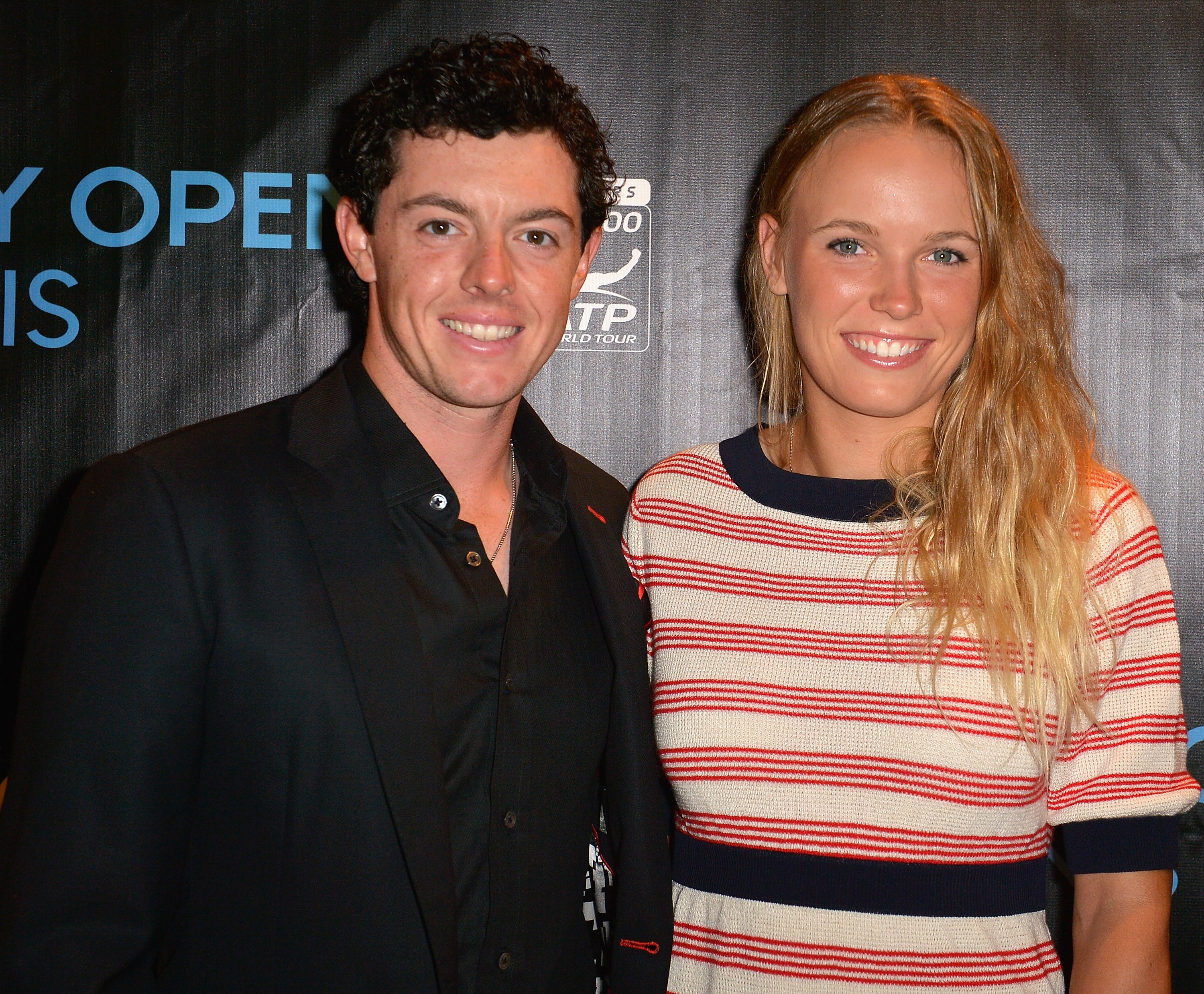Rory McIlroy in a dark suit standing next to Caroline Wozniacki in a striped top, both smiling for the camera at a professional event