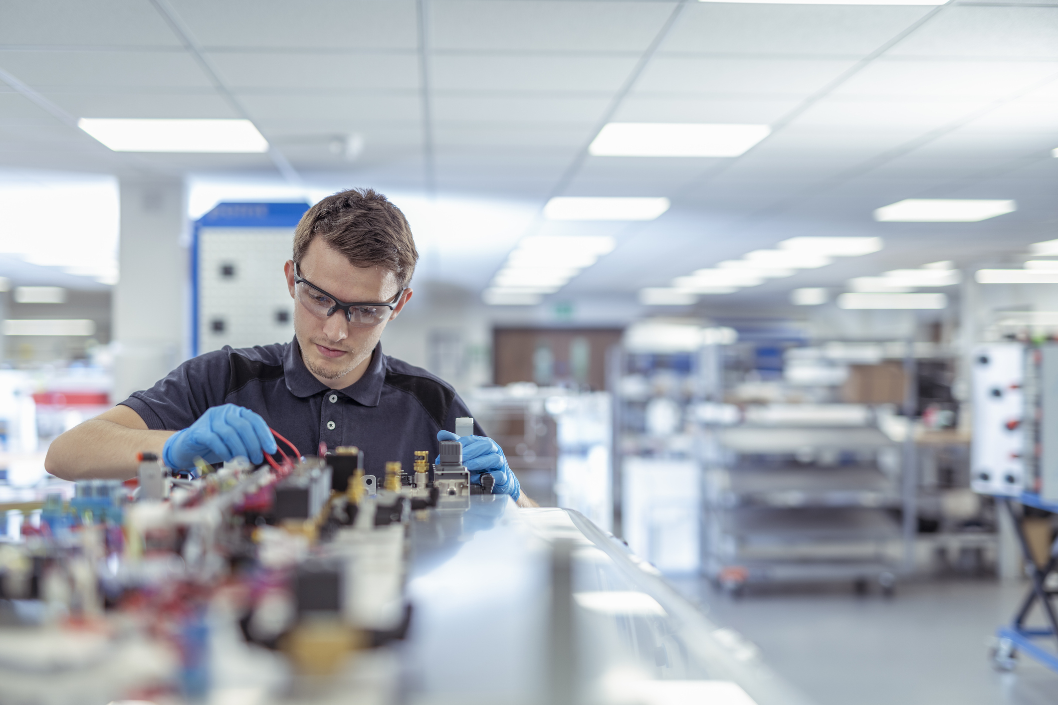 A man wearing safety glasses and gloves works with tools on an industrial assembly line in a well-lit factory setting