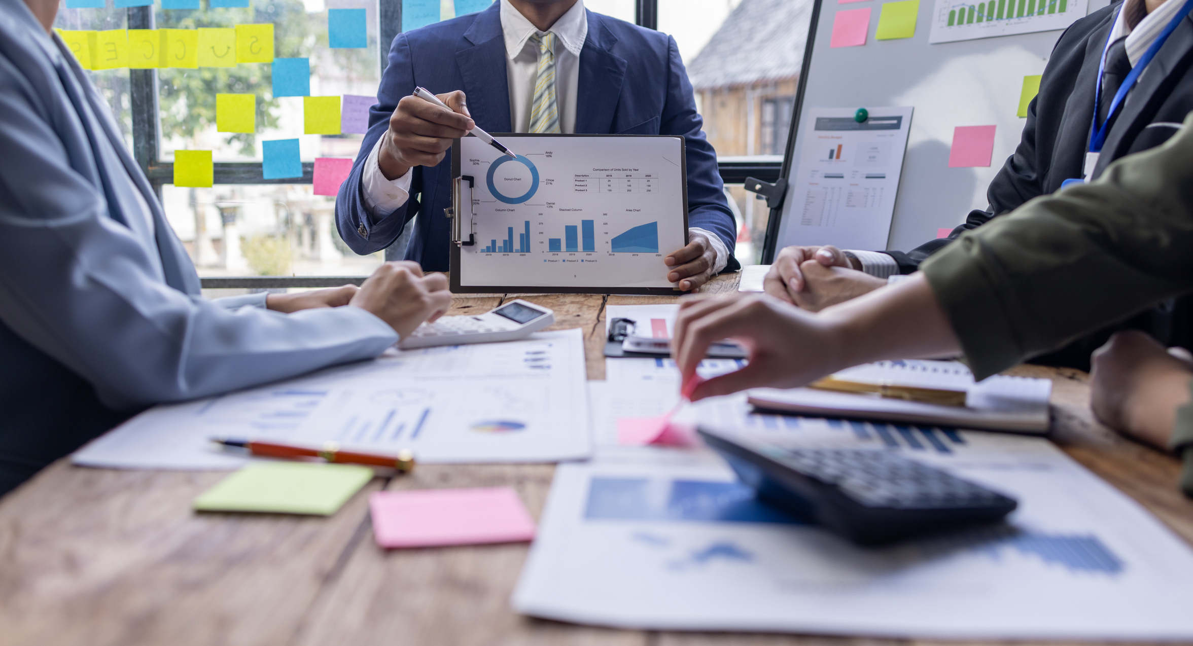 Business meeting with people examining charts and graphs. One person points to a pie chart on a document. Various documents and a calculator are on the table