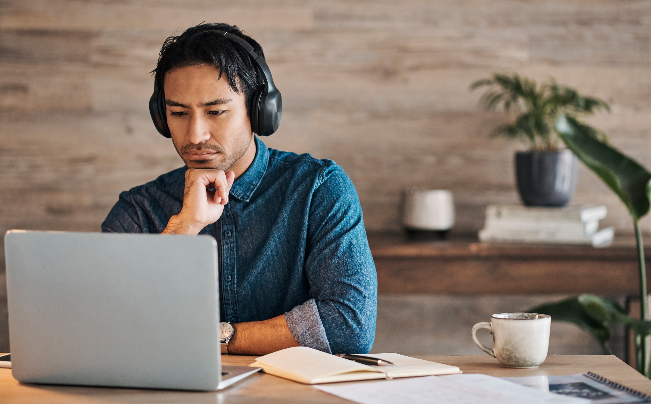 A man wearing headphones is working on a laptop at a desk with notebooks, a coffee cup, and a plant in the background