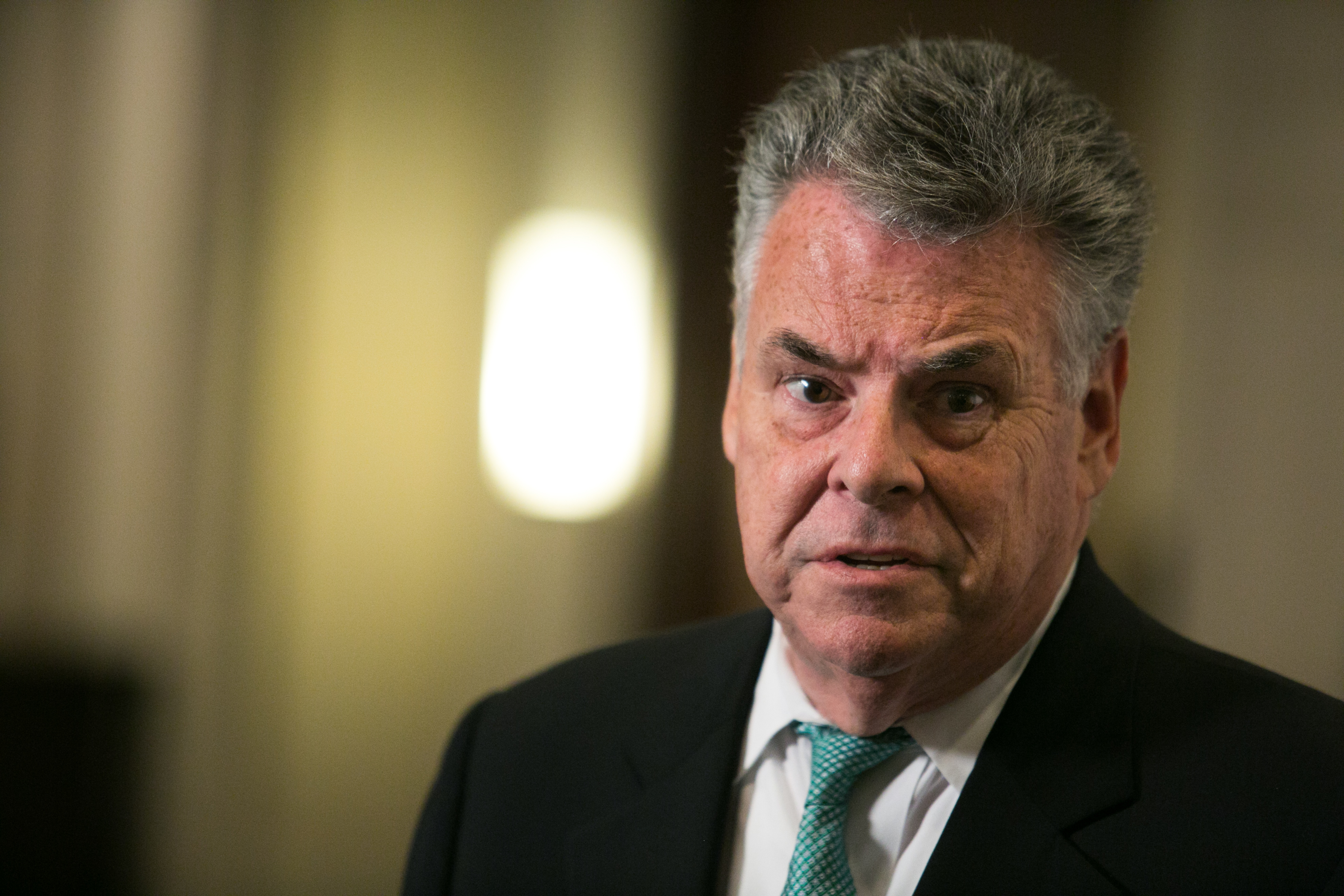 A man in a suit and tie, Peter King, looks directly at the camera with a neutral expression. The background is blurred